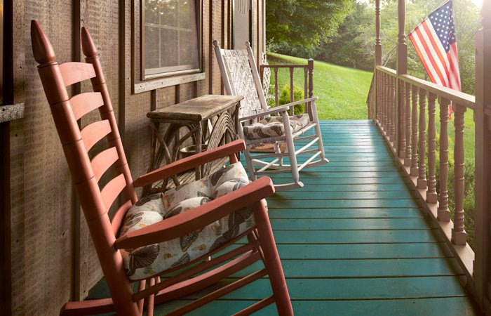 Two rocking chairs sit on the wooden porch of the Hawley house, alongside a small table and an American flag hanging from the railing.