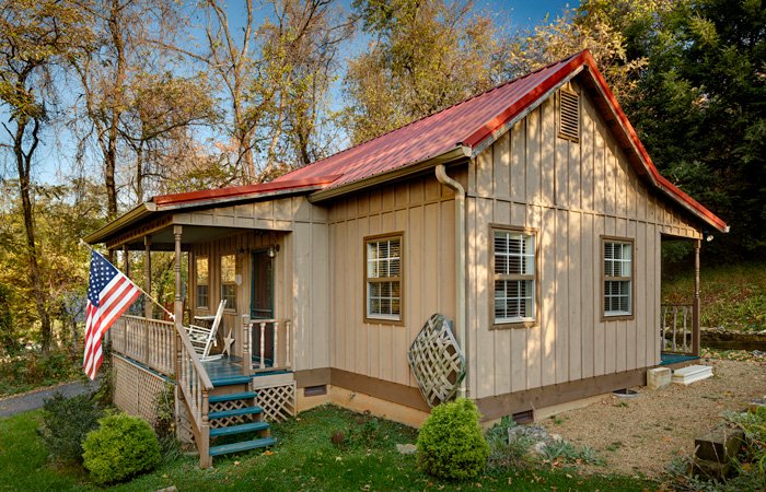 A tan board-and-batten Hawley house cottage with a red metal roof and a front porch featuring a rocking chair and an American flag.