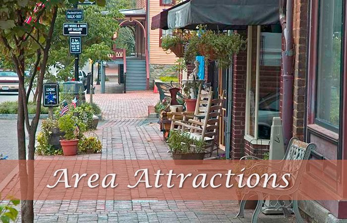 A brick sidewalk along the Hawley House storefront with wooden rocking chairs, potted plants, and street signs reading “Area Attractions.”